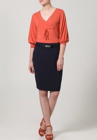 Haut cache-cœur couleur rouille avec un détail noué, assorti à une jupe crayon bleu marine foncé. Porté avec des sandales à talons hauts orange.