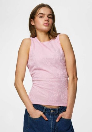 Ung kvinde iført en ærmeløs, pink tekstureret top og blå jeans, poserer med hænderne i lommerne mod en ensfarvet baggrund.