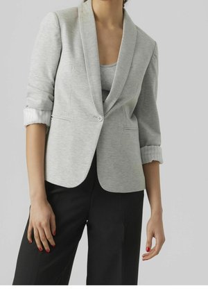 Vrouw draagt lichtgrijze blazer met één knoop over een bijpassende top en zwarte broek, met opgerolde mouwen en rode nagellak op handen.