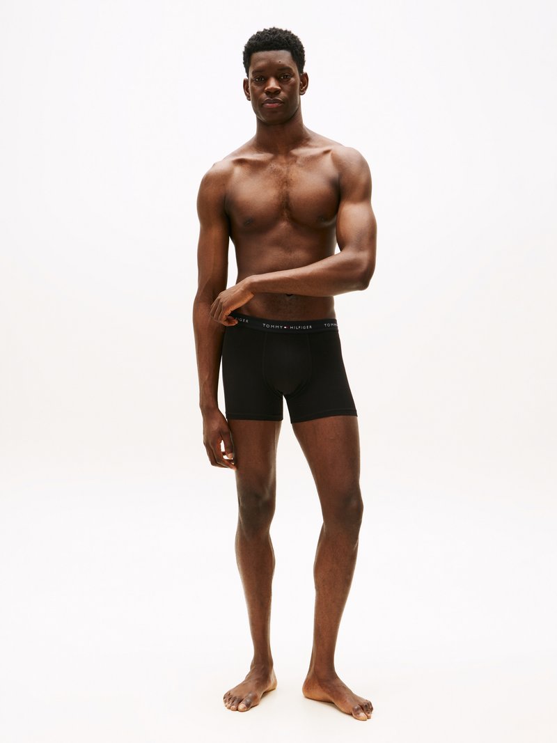 Fekete pamut boxer alsónadrág, testhez simuló kivitelben, elasztikus derékpánttal, amelyen a "Tommy Hilfiger" logó látható. A modell mezítláb áll, karjai lazán lelógatva.