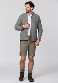 Grauer gestrickter Cardigan mit Reverskragen und Knopfverschluss, kombiniert mit hellbraunen Shorts mit gestickten Akzenten und Knöpfen. Braune Wanderschuhe.