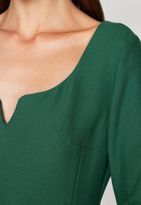 Gros plan sur l'épaule et la poitrine d'une femme portant un haut vert foncé avec une encolure unique à encoches et des manches longues.
