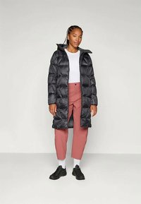 Svart boblejakke med langt design, høy krage og glidelås foran, kombinert med rosa bukser og svarte joggesko.