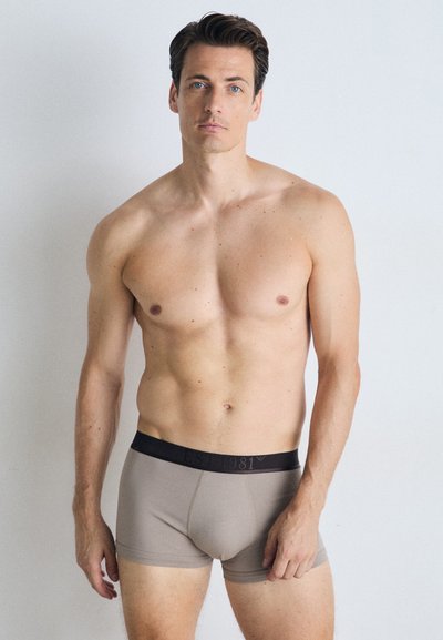 Beige boxer truser med svart elastisk midjebånd som har 'EST 1981' trykt i svart. Glatt tekstur, tettsittende design, midt på låret lengde.