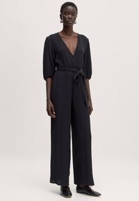 Zwarte jumpsuit met een diepe V-hals, gestructureerde stof, wijd uitlopende pijpen en een getailleerde taille met een strik. Heeft puff mouwen tot de elleboog en is in een effen kleur.