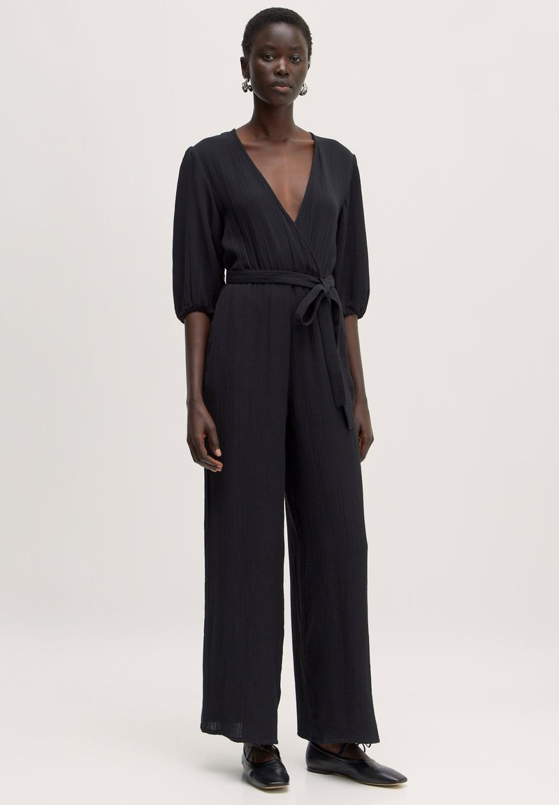 Zwarte jumpsuit met een diepe V-hals, gestructureerde stof, wijd uitlopende pijpen en een getailleerde taille met een strik. Heeft puff mouwen tot de elleboog en is in een effen kleur.