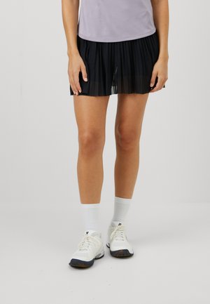 Svart plissert atletisk skjørt med gjennomsiktig lag, kombinert med en lys lilla topp og hvite sneakers med mørke detaljer.