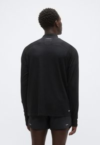 Camisola desportiva preta de manga comprida com bainha arredondada, textura macia e detalhe de logótipo refletor nas costas. Combinada com calções pretos.