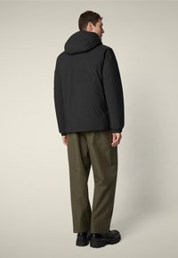 Giacca nera isolata con cappuccio, polsini elasticizzati e vestibilità casual, abbinata a pantaloni verde oliva e scarpe nere chunky.