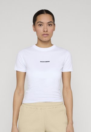 Femme aux cheveux foncés attachés porte un t-shirt blanc ajusté avec le texte « PEGADOR » et un pantalon beige, debout devant un fond neutre.