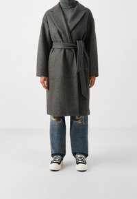 Grå ullblandad kappa med bälte i midjan, slagkrage och långa ärmar, tillsammans med slitna blå jeans och svartvita sneakers.