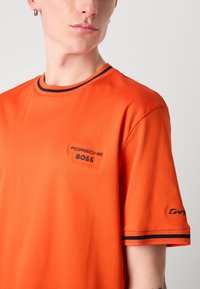 Oranžinė medvilninė marškinė su juodu apykakle, dryžuotomis detalėmis ir siuvinėtu užrašu "PORSCHE BOSS" ant krūtinės bei "targa" ant rankovės.
