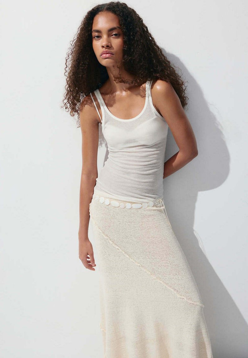 Femme aux cheveux bouclés portant un débardeur blanc et une longue jupe beige, debout devant un fond clair uni avec ombre.