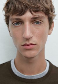 Nahaufnahme eines Porträts einer Person mit hellbraunen Haaren und grünen Augen, die ein dunkelbraunes Hemd über einem grauen Rundhalsausschnitt-T-Shirt trägt.