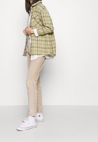 Žena oblečená v béžových manšestrových kalhotách, bílých vysokých teniskách, bílém tričku a zelené kostkované bundě, stojící na bílém pozadí.