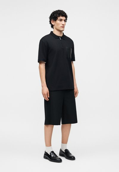 Νέος άνδρας στέκεται φορώντας μαύρο πόλο με κοντά μανίκια, μαύρα σορτς μέχρι το γόνατο, λευκές κάλτσες και μαύρα δερμάτινα μοκασίνια σε απλό φόντο.