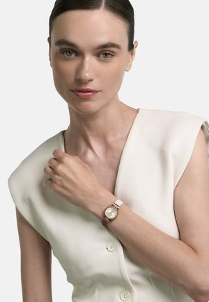 Femeie cu păr dat pe spate poartă un top alb fără mâneci și un ceas de mână aurit cu o curea bicoloră în roz și burgund.