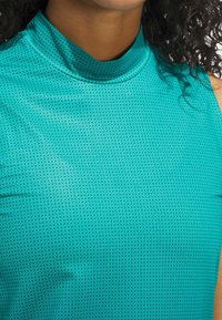 Turkos topp med hög krage i texturerat, andningsbart tyg med små svarta prickar över hela. Slät yta med ärmlös design.