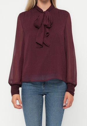 Femme portant un chemisier bordeaux transparent à manches longues avec un nœud au col, associé à un jean bleu.