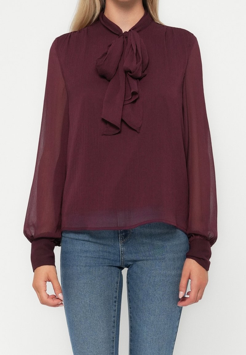 Femme portant un chemisier bordeaux transparent à manches longues avec un nœud au col, associé à un jean bleu.