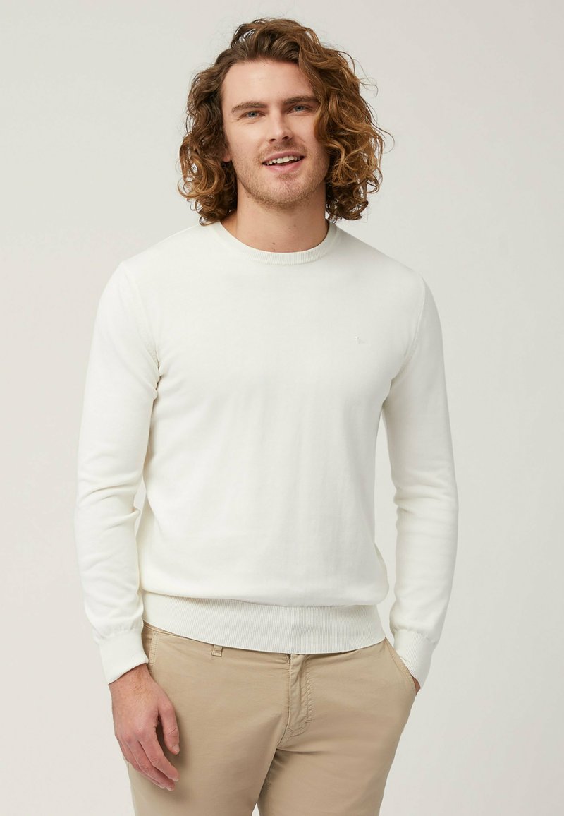 Maglione bianco a maniche lunghe con scollatura rotonda e polsini a coste. Design aderente con un sottile dettaglio del logo sul petto. Indossato con pantaloni beige chiaro.