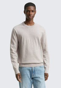 Mann steht mit dem Gesicht nach vorne, trägt einen hellbeigen Pullover mit Rundhalsausschnitt und hellblaue Jeans vor einem schlichten, hellen Hintergrund.