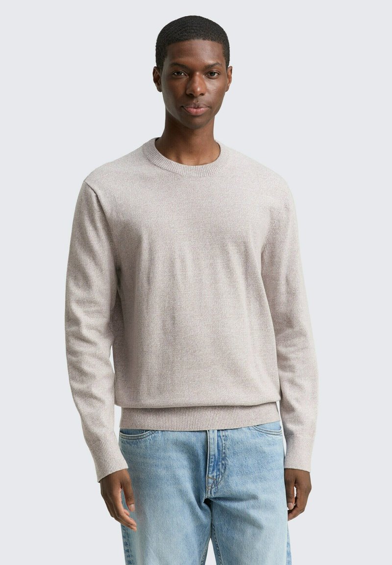 Mann steht mit dem Gesicht nach vorne, trägt einen hellbeigen Pullover mit Rundhalsausschnitt und hellblaue Jeans vor einem schlichten, hellen Hintergrund.