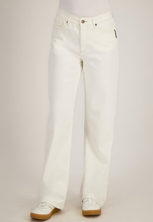 Vrouw draagt off-white rechte jeans en witte sneakers met gumzolen, staande tegen een effen lichte achtergrond.