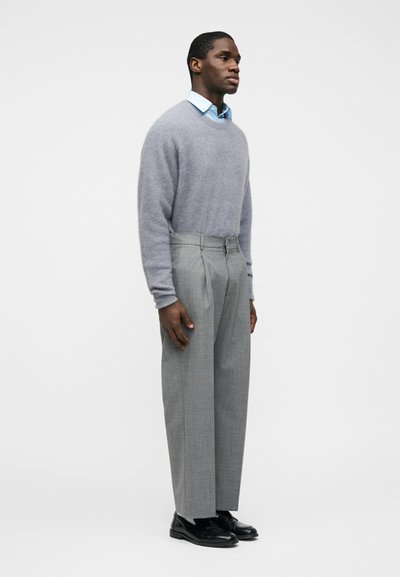 Άνδρας όρθιος με τα χέρια δίπλα φοράει ανοιχτό γαλάζιο πουκάμισο με γιακά, γκρι αφράτο πουλόβερ, γκρι πιετέ παντελόνι και μαύρα παπούτσια κοστουμιού.