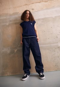 Navy sleeveless top with a ribbed neckline, paired with loose-fitting dark denim jeans and black sneakers with white accents. Plain background.