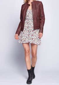 Mujer con chaqueta de cuero burdeos, vestido floral blanco y botas negras con cordones hasta el tobillo, de pie contra un fondo claro y liso.