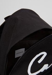 Czarny plecak z zamykaną komorą. Posiada duże białe logo oraz etykietę pielęgnacyjną w środku, która ukazuje gładką teksturę materiału.