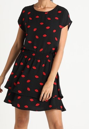 Femme portant une robe noire à manches courtes avec des imprimés de lèvres rouges et une taille élastique, debout devant un fond uni.