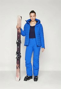 Μπλε μπουφάν με κουκούλα, ταιριαστά μπλε παντελόνια σκι και μαύρες μπότες σκι; κρατώντας ένα ανοιχτό καφέ σκι με μαύρους δακτυλίους.