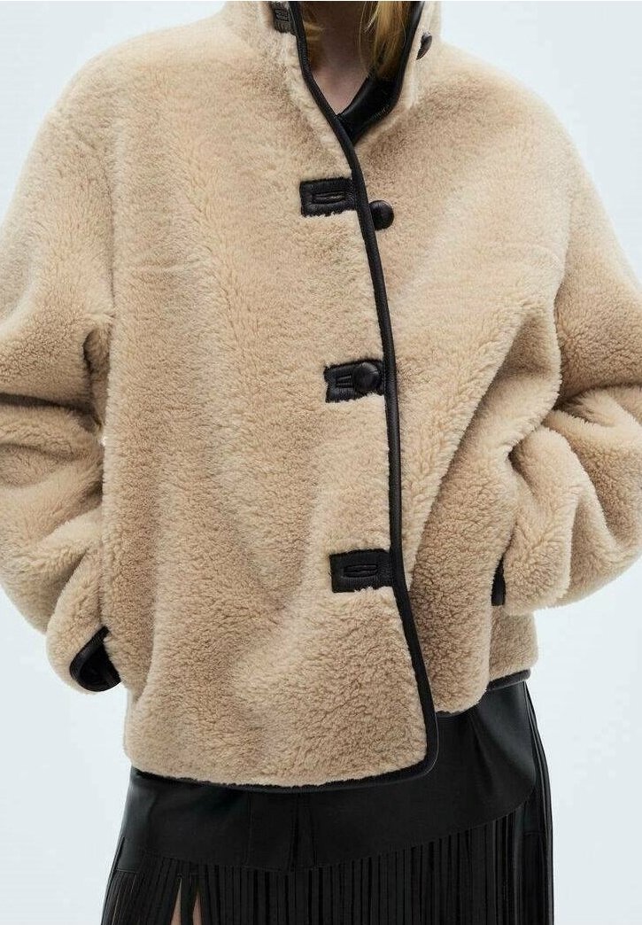 Veste beige duveteuse avec bordure noire et boutons à bascule portée sur une jupe noire plissée, mains dans les poches de la veste.