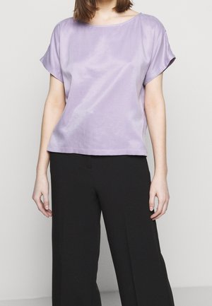 Personne portant un haut ample lilas à manches courtes et un pantalon noir à jambes larges, debout devant un fond clair uni.