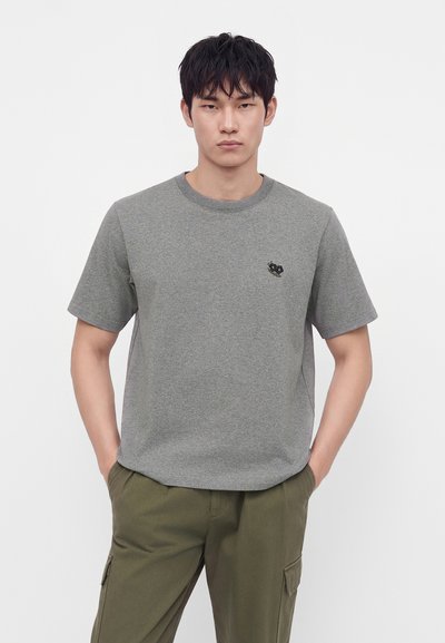Un jeune homme est debout, tourné vers l'avant, portant un t-shirt gris à manches courtes avec un petit emblème de fleur noire et un pantalon cargo vert olive.