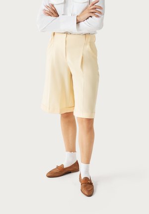 Persona che indossa pantaloncini beige fino al ginocchio, calzini bianchi, mocassini marroni e una camicia bianca a maniche lunghe con le braccia incrociate, in piedi su uno sfondo chiaro.