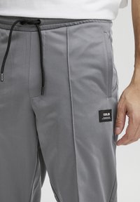 Pantalones de atletismo grises fabricados con un tejido suave, con una cintura elástica y un cordón negro, además de una pequeña etiqueta de tela negra en el lateral.