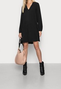 Vestido negro de manga larga con escote en V y diseño en capas; combinado con un bolso de mano rosa claro y botas de tobillo negras con tacón. Tejido suave.
