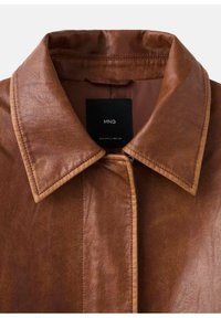 Veste en cuir marron avec col pointu et détails de surpiqûres visibles. Étiquette noire avec le nom de la marque à l'intérieur. Texture lisse.