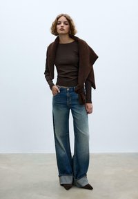 Top marrone a maniche lunghe, jeans larghi in denim con tonalità blu sbiadite, tacchi arrotondati marroni e uno scialle marrone drappeggiato sulle spalle.