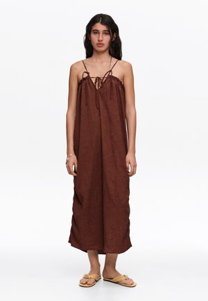 Joven mujer que lleva un vestido midi marrón suelto y sin mangas con tirantes finos y sandalias planas beige, de pie frente a un fondo blanco liso.