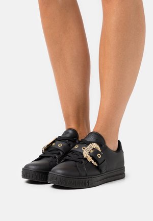 Sorte lave sneakers med guldkæde spænde detaljer, sorte snørebånd og præget brandnavn på sålen, båret på bare fødder.