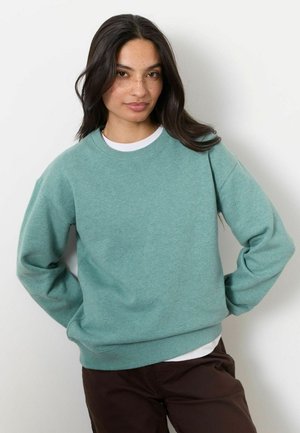 Kvinde med langt mørkt hår og fregner iført en grøn sweatshirt over en hvid skjorte og mørke bukser, stående med hænderne på hoften.