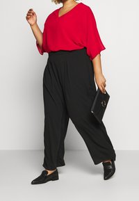 Blouse rouge à manches courtes avec col en V, associée à un pantalon noir à jambes larges. Pochette noire et chaussures à enfiler complètent la tenue.