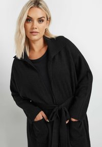 Mustasta neulotusta neuletakista, jossa on shaala-kaulus, pitkät hihat ja nauhavyötärö. Siinä on ribbiä hihansuut ja kaksi sivutaskua. Pehmeä rakenne.