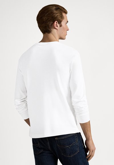 Hosszú ujjú fehér póló kerek nyakkivágással, sima anyagból és laza szabással, sötétkék farmernadrággal, mely hátsó zsebekkel rendelkezik.