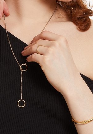Femme portant un haut noir, tenant un collier lariat en or avec des pendentifs circulaires ; elle porte également une bague en or et un bracelet à la main gauche.