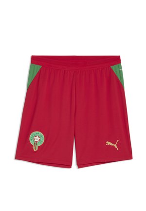 Røde atletikshorts med grønne sidelommer, med et gyldent Puma-logo og en grøn cirkulær embleme med arabisk tekst på venstre ben.